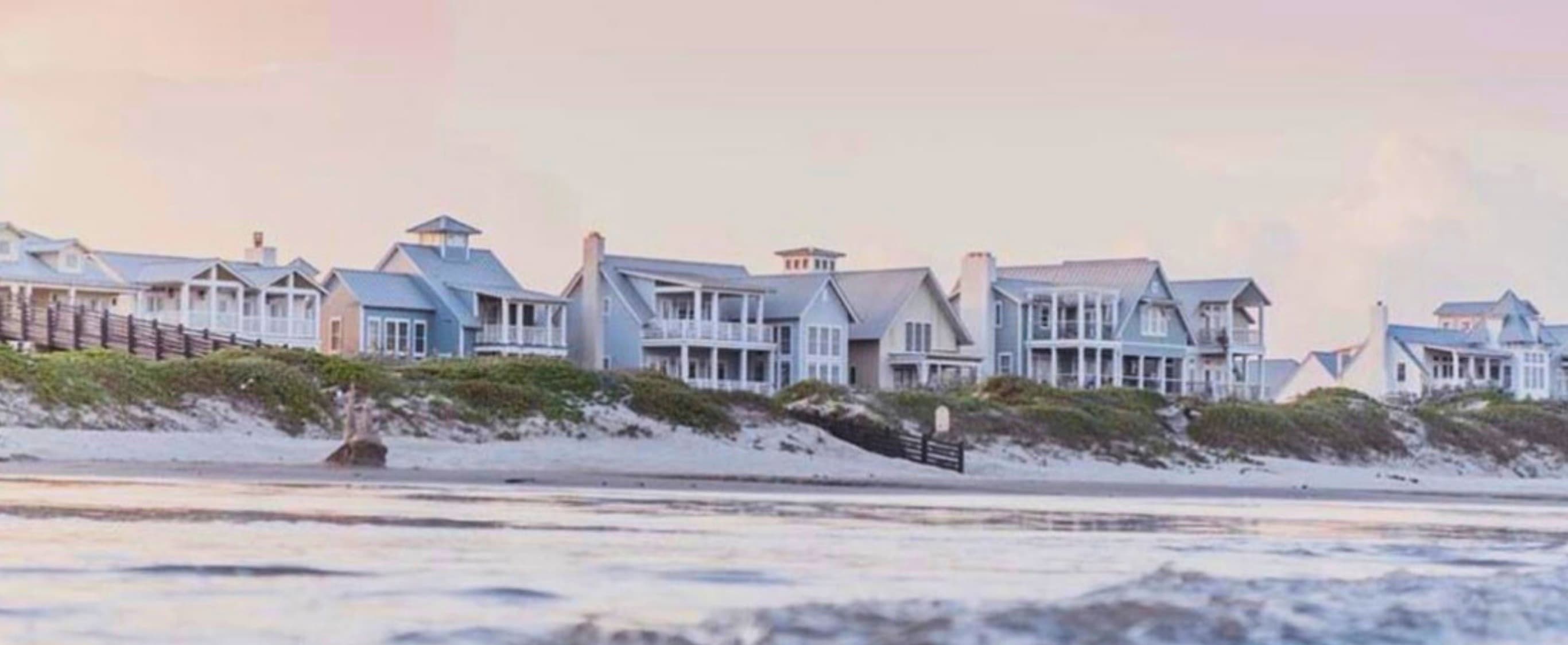 Cinnamon Shore — pastel sunset over the coastal community, beach houses lining the dunes with surf in the foreground