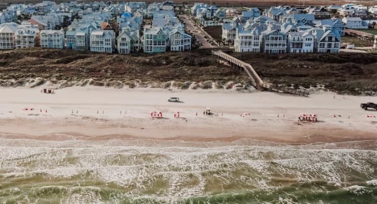 Cinnamon Shore aerial — pastel beach houses lining the dunes with surf in the foreground