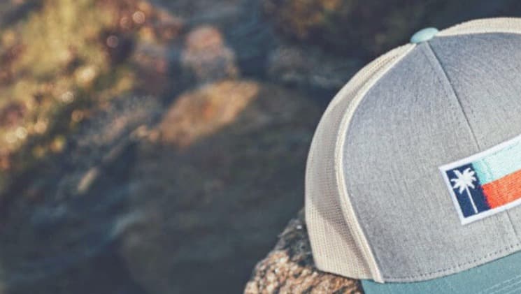 The Palm Republic — heather-grey trucker cap with Port Aransas flag patch sitting on coastal rocks