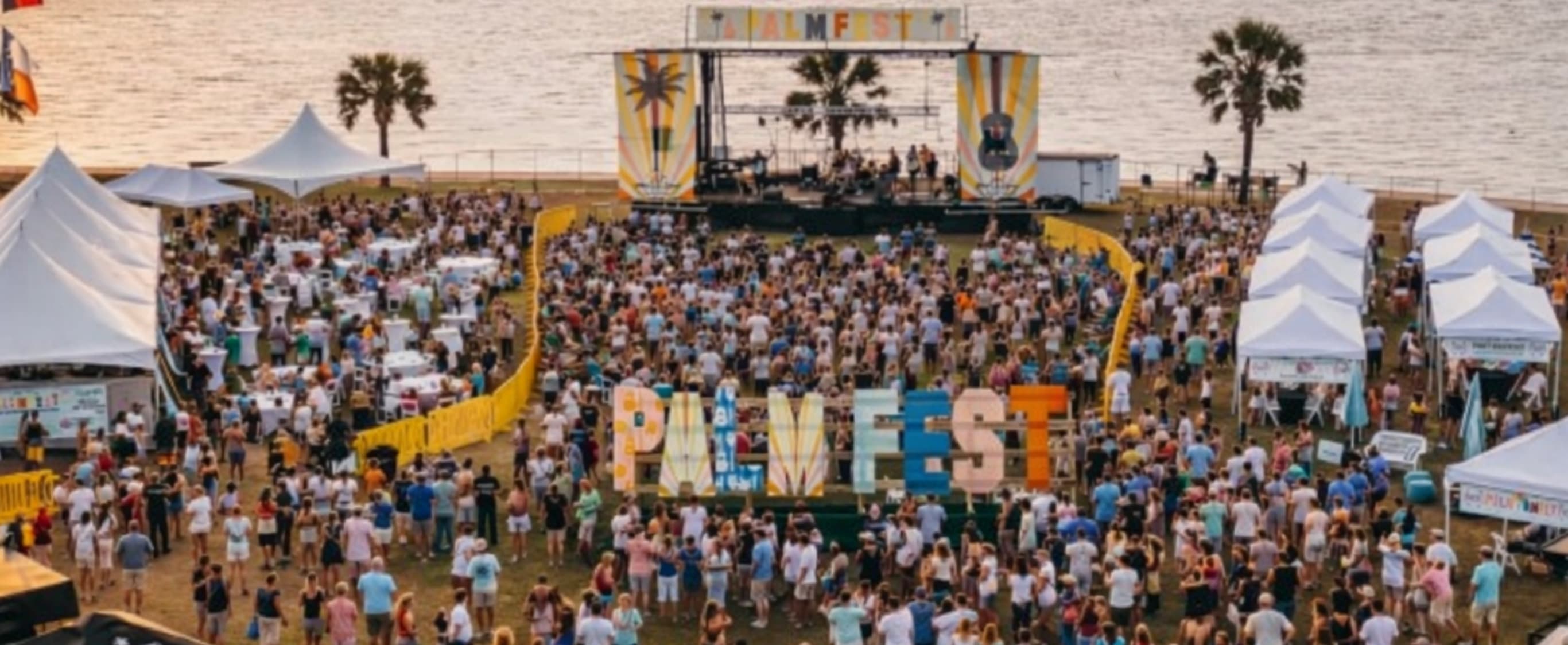 PALMFEST aerial — main stage with PALMFEST banner, crowd around the giant PALMFEST letter installation, white tents, palm trees, and the Gulf